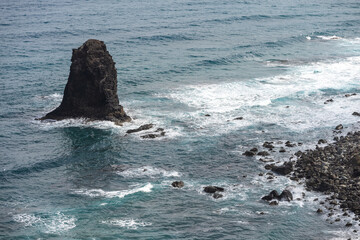Dramatic volcanic rock formation rising from turquoise ocean waves and rocky shoreline near Benijo beach, Tenerife, Canary Islands, Spain.