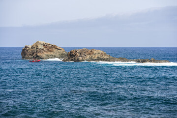Rescue motor boat in red color maneuvering through ocean waves next to prominent volcanic rock formation at Roque de las Bodegas, Tenerife, Canary Islands, Spain.