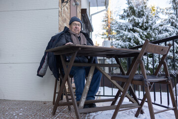 Senior male sitting outside house reading print media during breakfast in warm clothes. Quiet moment on lifestyle scene.