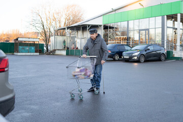 Elderly man in outdoor clothing standing on parking lot with shopping cart full of groceries near personal vehicle