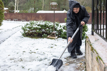 Elderly man in dark winter clothing clearing snow with shovel on paved yard area emphasizing seasonal outdoor work
