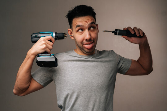 Portrait of fooling young man playing funny joke, using large cordless drill as earphone and small electric screwdriver as portable music player, having fun and making silly face.