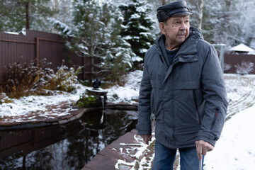 Elderly man walking with cane on paved yard after snowfall wearing winter jacket and hat outdoors calm winter environment.