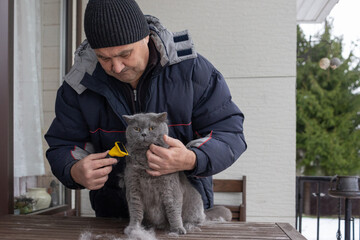 Elderly man in warm winter clothing gently grooming gray cat with brush emphasizing care responsibility and human animal bond.