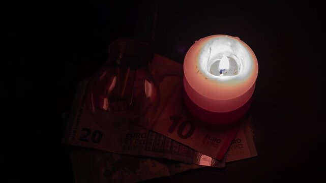 A burning candle, a dead cell phone, and a disconnected lamp lie on top of banknotes. Power outages due to the war in Ukraine.