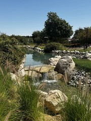 Landscaped water feature with rocks, grass, and trees under a clear sky.