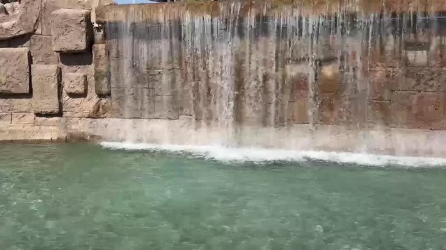 A cascading waterfall flows into a pool of water, with a stone structure surrounding it. Ripples are visible on the water's surface.