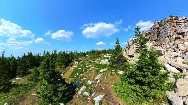 A wide-angle shot of a picturesque hiking trail leading up a rocky, forested mountain slope under a bright blue sky with white, fluffy clouds. The landscape features dense green trees, large stone bou