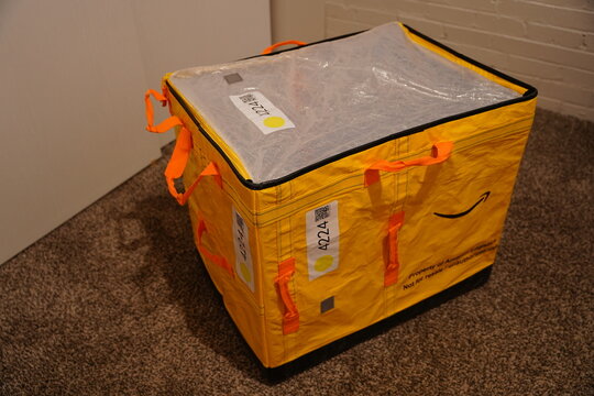 Yellow Amazon Logistics delivery tote bag with smile logo sitting on a carpeted floor in a home or warehouse setting for editorial use.