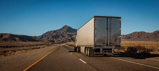 Following behind a white truck on a two lane highway in the desert with a rocky hill in the distance. The image is from the I-15 in Nevada