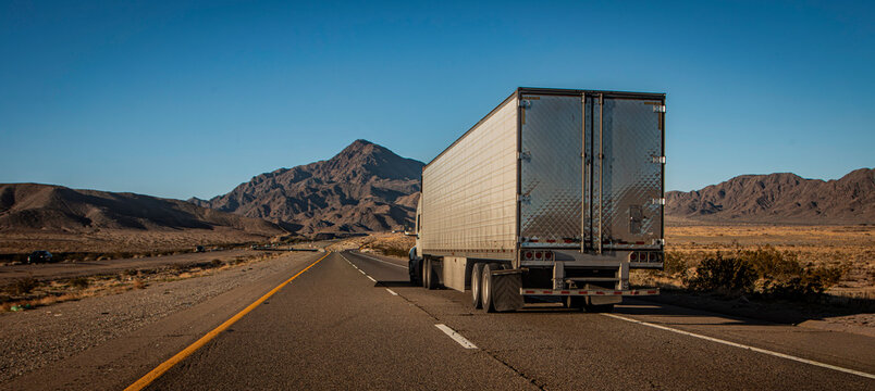 Following behind a white truck on a two lane highway in the desert with a rocky hill in the distance. The image is from the I-15 in Nevada