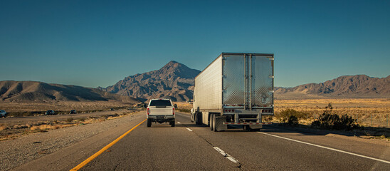 Following behind a white truck and trailer and a white pick up truck  on a two lane highway in the desert . The image is from the I-15 in Nevada