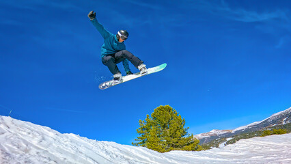 LOW ANGLE VIEW: Male snowboarder in blue jacket makes a steezy melon grab while jumping on a fun feature in snowpark. Freestyle rider enjoys doing park laps at sunny ski resort in snowy Austrian Alps.