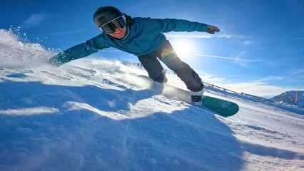 CLOSE UP, LENS FLARE, LOW ANGLE VIEW: Smiling snowboarder in blue jacket drags his hand and sprays snow while carving on sunny ski slope. Winter fun and authentic snowboard action in Austrian Alps. © helivideo