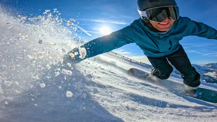 CLOSE UP, LENS FLARE, LOW ANGLE VIEW: Cheerful male snowboarder carves on sunny ski slope. While he drags his hand on snow, a cloud of white ice crystals spray through air. Winter fun in the Alps. © helivideo