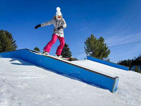 Freestyle snowboarder in pink pants slides a blue box rail in a terrain park. Young woman is doing a stylish boardslide trick during a fun lap through snowpark at Austrian ski resort on a sunny day.