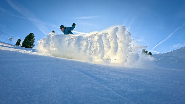 LENS FLARE, LOW ANGLE VIEW, SILHOUETTE: Snowboarder sends a cloud of snow towards camera as he makes a turn while riding down the ski slope. Fun snowboarding at ski resort in the snowy Austrian Alps.