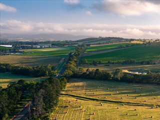 Golden hour illuminates a vast agricultural landscape featuring hay bales, green fields, a winding road, and distant misty hills. © Mujie