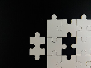 Fototapeta premium Minimalist top-down view of a white jigsaw puzzle on a black background. One piece is separated, symbolizing the final step, solution, and business completion.