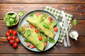 Tasty spinach crepes with tomatoes, onion, sour cream and leaves on wooden table, flat lay