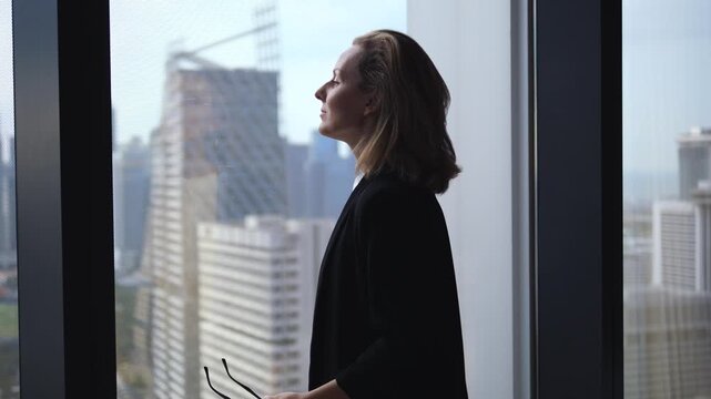 Side view of a stressed businesswoman taking off her glasses and looking out the window at the cityscape, thinking about her business strategy and feeling anxious and overwhelmed
