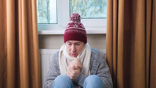 Freezing man in hat and scarf sitting by cold radiator, rubbing hands to warm up at home during winter. High quality 4k footage