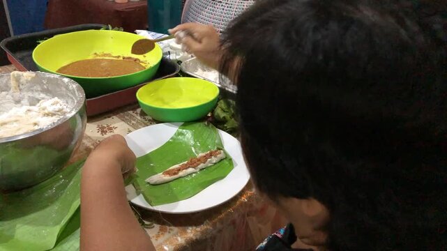 Flavored glutinous sticky rice is rolled and wrapped in a banana leaf to create homemade steamed rice cakes on a cluttered work table by an elderly woman.