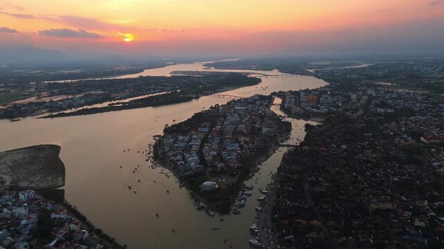 Breathtaking high-angle drone shot over Hoi An, Vietnam at twilight. Watch the traditional wooden boats glide along the Thu Bon River as the sunset paints the UNESCO Ancient Town in warm golden hues