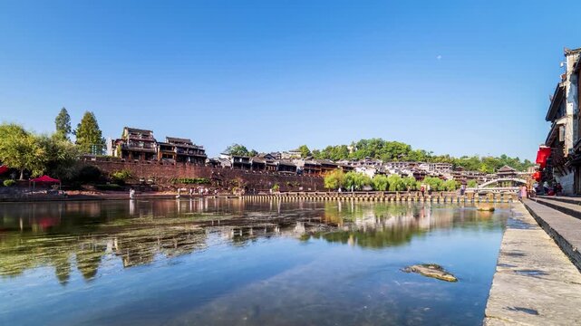 slow-shutter time lapse of Fenghuang Ancient Town's Tuo River with stone stepping bridge, traditional buildings, and bustling tourists motion blur under a clear blue sky, Hunan Province, China
