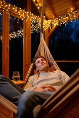 A young man lies back smiling in a hammock on a cozy wooden deck at night, surrounded by twinkling fairy lights and enjoying a glass of wine nearby. Generative AI.