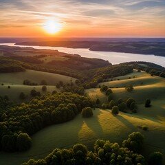 Aerial view of a rolling green landscape at sunset, with a large lake and distant hills. The sky is painted with warm orange hues
