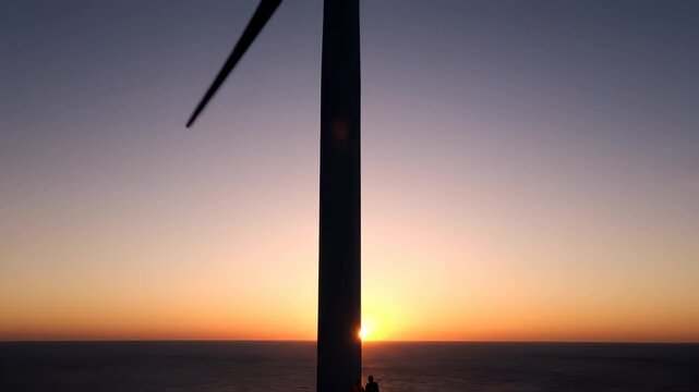 Person stands beside wind turbine at sunset over ocean. Turbine blade frames sunset on sea horizon. Wind energy tower shows person at sunset. Ocean turbine with sunset sky and person below.