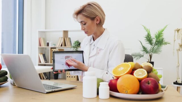 A female dietitian explains the digestive system using a tablet, surrounded by fruits and vegetables, indicating a focus on healthy eating.