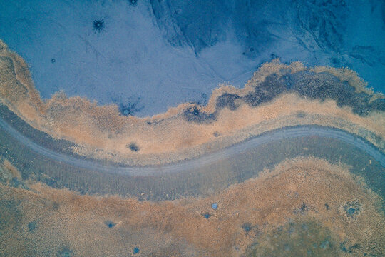 Curved dirt road separating toxic blue sludge and dry grass, surreal industrial pollution texture and landscape background, aerial top view drone shot