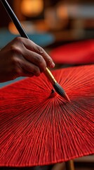 Close-up of a hand delicately painting a vibrant red umbrella with thin brush, capturing traditional craftsmanship and intricate detail in the workshop setting.Hand