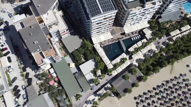 Drone view of a popular beachfront with rows of umbrellas, a wide promenade, and seaside buildings during a bright summer day.