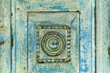 Architectural fragment in Ermoupolis, Syros, Greece, showing a weathered wooden panel with concentric circular carvings set within a square frame. 