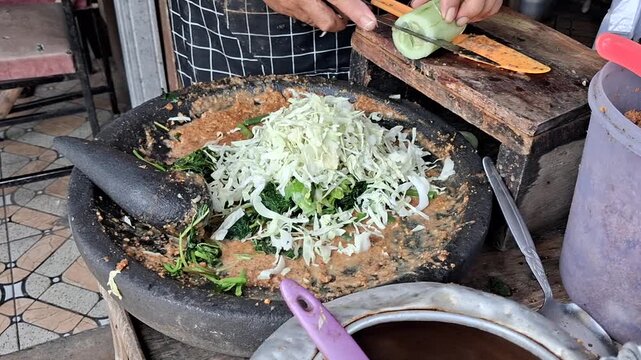 ​Footage of making Lotek, a traditional Indonesian salad featuring steamed vegetables and sliced fresh cucumber, mixed with savory peanut sauce in a stone mortar.