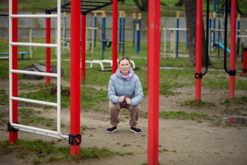 Focused woman in a blue jacket squats at an outdoor gym, showcasing fitness and an active lifestyle. Her urban workout is set amidst playground equipment and natural surroundings.