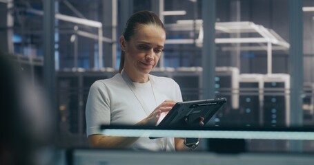 Woman, IT Specialist Stands and Uses Portable Tablet Device to Check Real Time Performance Metrics in High Security Server Hall. Concept of System Monitoring, on Site Expertise, and Data Analysis.
