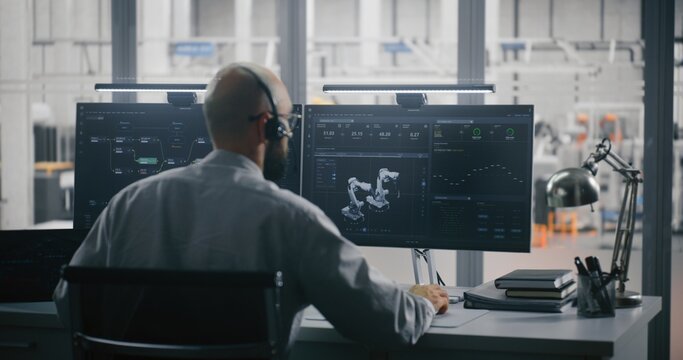 Engineer on Headset Controlling 3D Automation Model. Man Working Intensely at Two Monitors. Screens Display 3D Robot Model and Metrics, Overlooking Automated Factory Floor. Remote Robotics Management. - Powered by Adobe