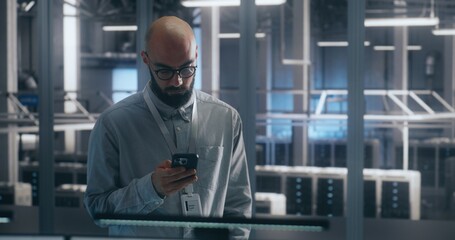 Bearded Male IT Engineer in Glasses Stands and Uses Smartphone to Check Critical Notifications or System Status Large Scale of Data Center. Emphasizes Mobile Management and Operational Urgency.