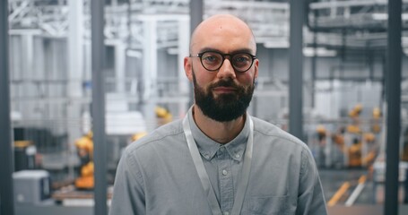 Dedicated Male Technical Specialist Poses Confidently in Front of Automated Industrial Plant Containing Machinery. Concept of Role in Managing Robotics and Advanced Manufacturing Processes. Portrait.