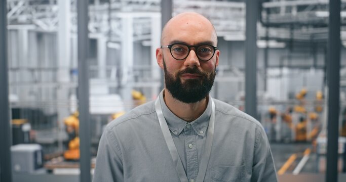 Dedicated Male Technical Specialist Poses Confidently in Front of Automated Industrial Plant Containing Machinery. Concept of Role in Managing Robotics and Advanced Manufacturing Processes. Portrait.