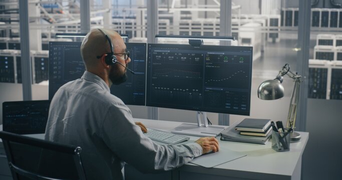 Specialist Coordinating Data Center Operations Remotely. IT Specialist Speaking on Headset, Actively Typing at Desk With Dual Monitors in High Security Data Center. Concept of Focused Data Analysis.