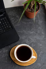 A cup of black coffee on a wooden coaster next to a laptop and a potted plant. The scene is set on a dark textured surface.