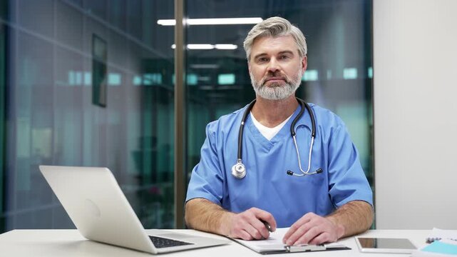 Portrait of serious mature doctor in blue coat works in office sitting at desk at workplace in hospital clinic. Confident male professional medical worker physician writing paperwork looking at camera