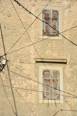 A weathered stone facade of an old mediterranean building features two windows with closed rustic...