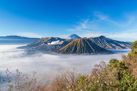 Morning view, Mount Bromo, East Java, Indonesia. Smoke rising from one of the active volcanoes, Fog covering the surrounding desert. Blue sky above. 
