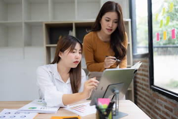 Two asian businesswoman using digital tablet and talking about business with analyzing financial charts in the modern office.